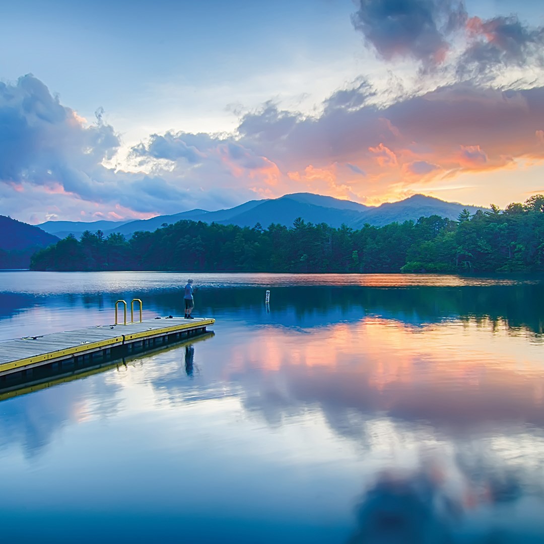 A person stands on a wooden dock on calm Lake Santeetlah at sunset, with mountains and a colorful sky mirrored in the still water.