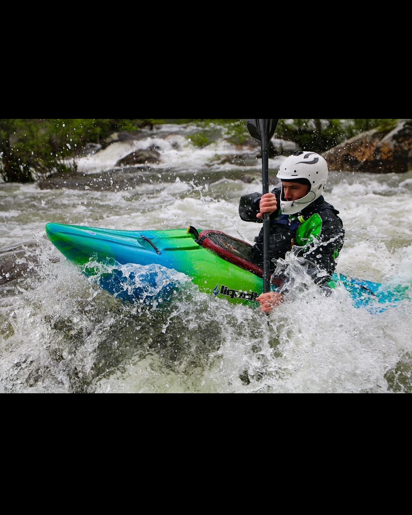 A helmeted kayaker paddles a green kayak through churning whitewater rapids, spray flying around the boat.
